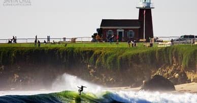 Surfer carving wave near coastal lighthouse on sunny bluff