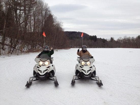Two snowmobiles side-by-side on frozen lake trail in winter woods