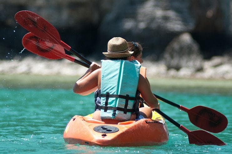 Tandem kayakers paddling on calm teal bay near rocky coast
