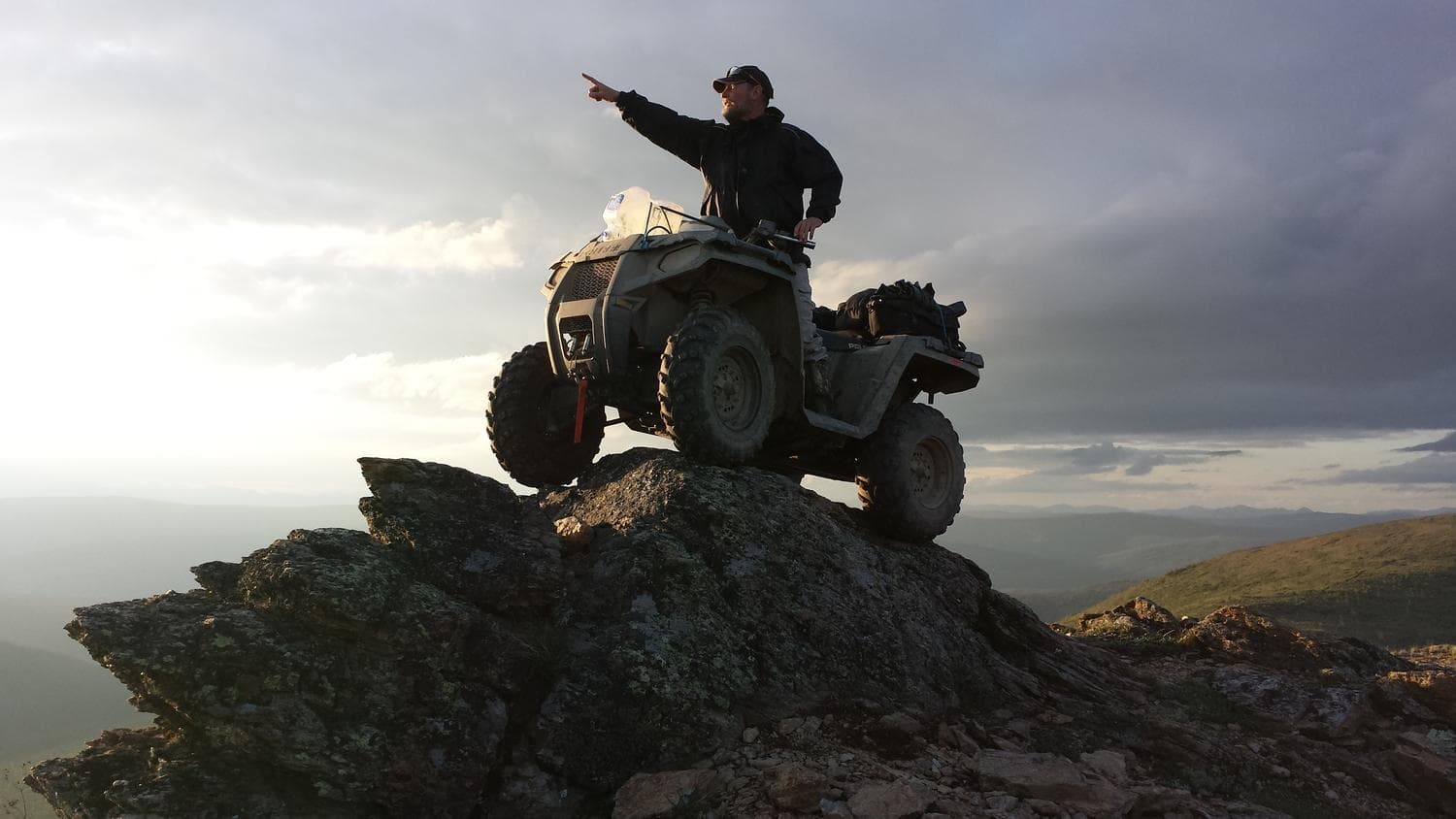 ATV rider scouting coastal tundra from rocky summit at sunset