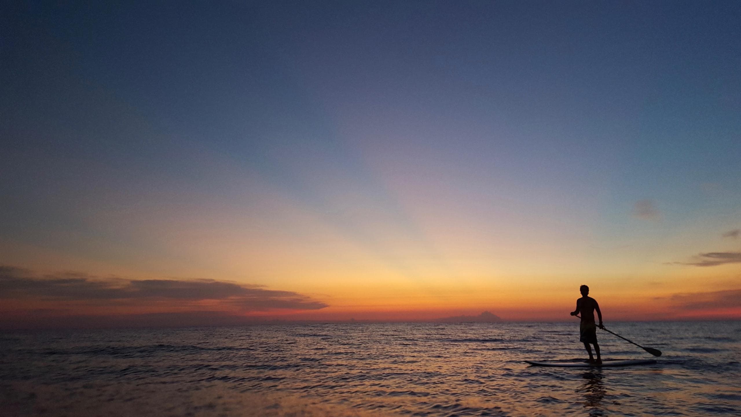 Paddleboarder silhouetted at sunset over ocean with colorful sky