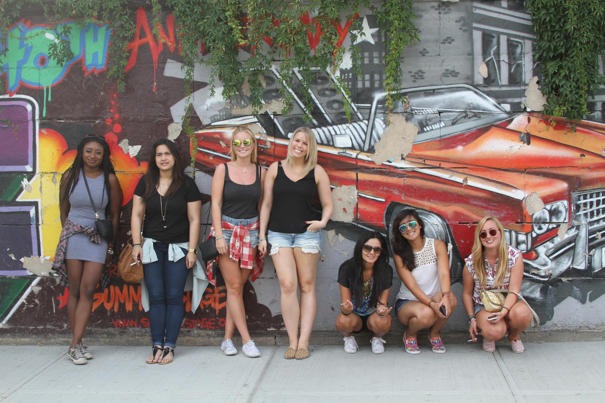 Group posing by colorful classic car mural on city sidewalk