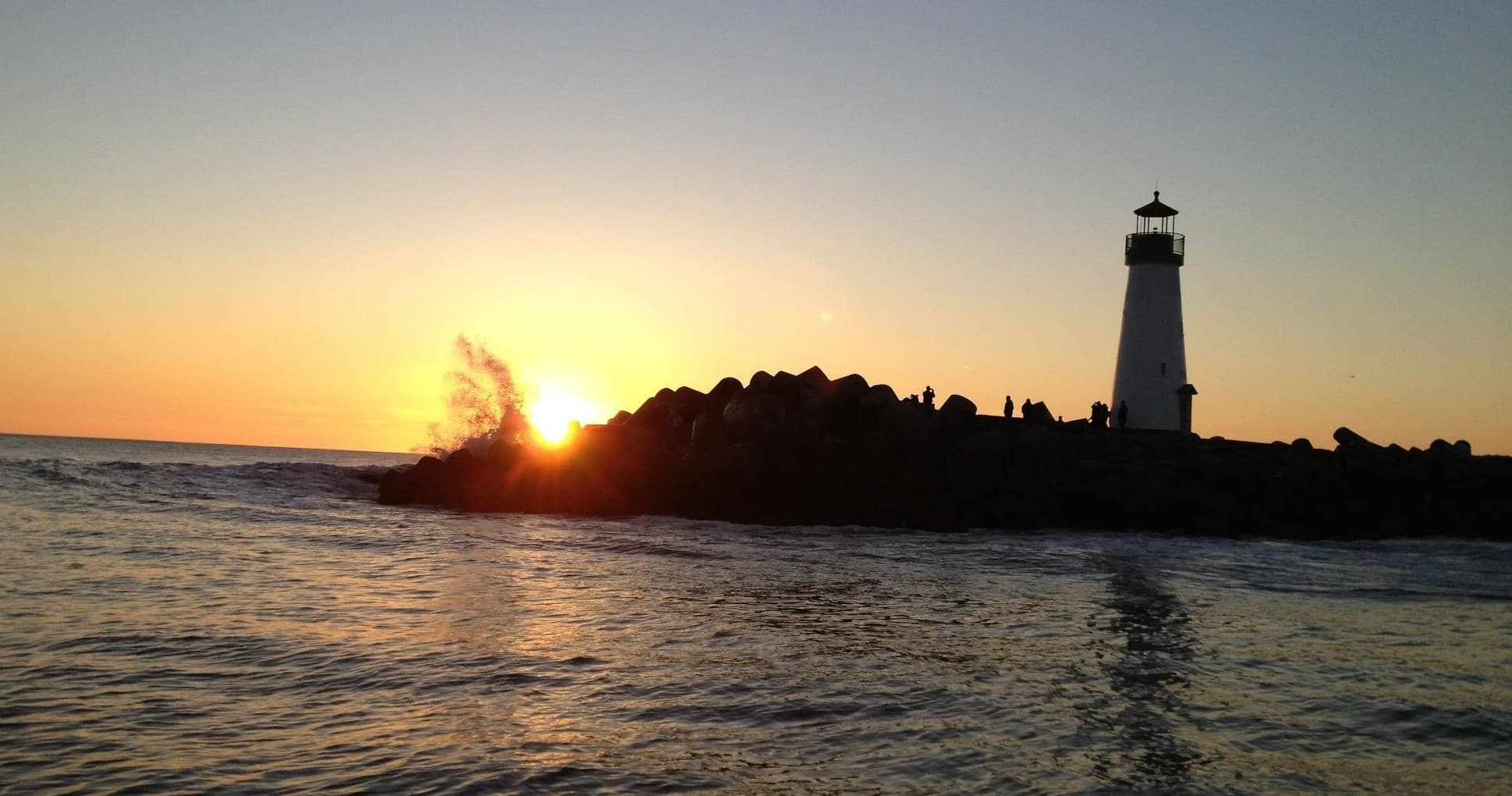 Lighthouse silhouetted at sunset over rocky breakwater in California