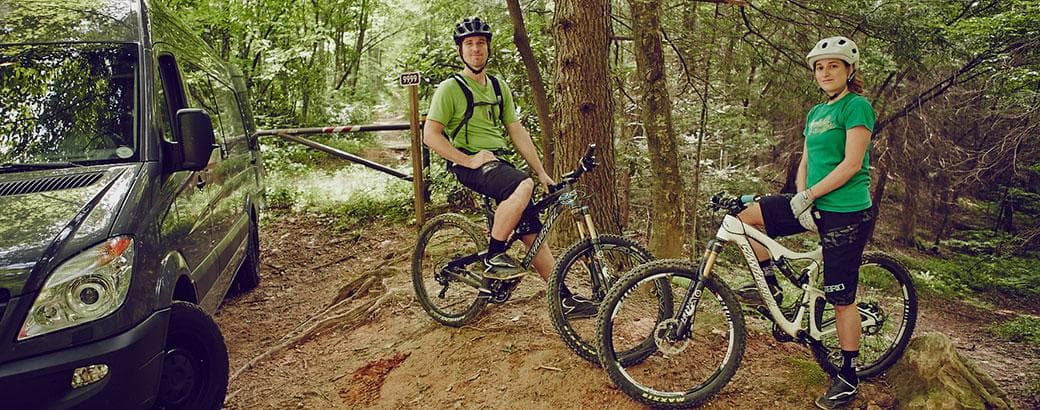 Two cyclists pausing on a shaded forest trail beside a vehicle