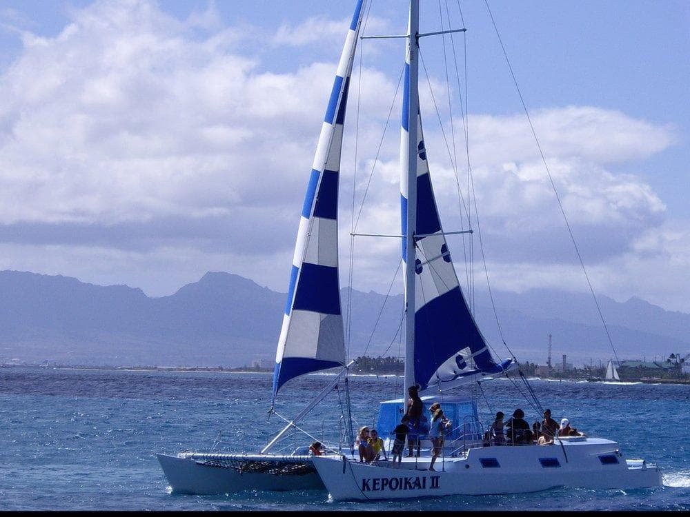Catamaran sailing with blue-striped sails off a mountainous coast